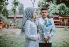 A Muslim couple in traditional attire shares a moment outdoors with a floral bouquet.