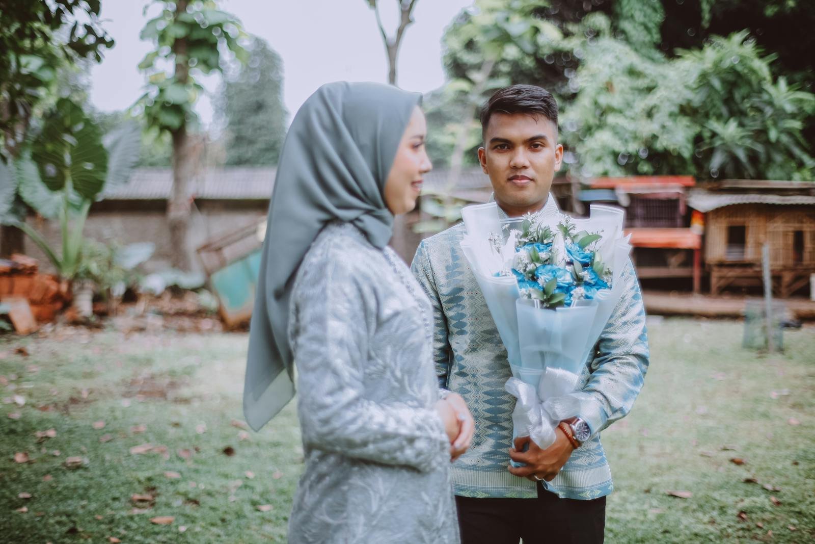 A Muslim couple in traditional attire shares a moment outdoors with a floral bouquet.